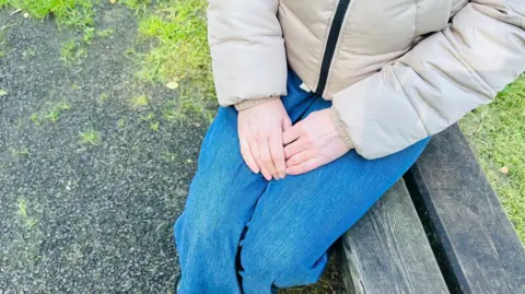 A woman's hands clasped together. She wears jeans and a beige puffer coat