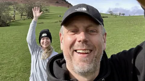 Ed Jenkins Ed taking a selfie with Helen in the background, waving. They are both smiling and wearing hoodies. Ed wears a black baseball cap and Helen wears a black beanie.