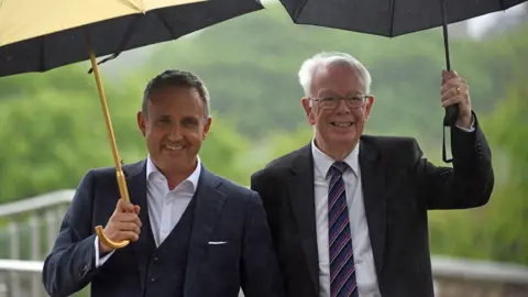 PA Media Alex Cole-Hamilton, a smiling man with greying hair wearing a blue three-piece suit and white open-neck shirt, is standing under an umbrella, sheltering from heavy rain. He is smiling. To his left and on the right of the picture is Jim Wallace is dark grey suit with white shirt and purple and white striped tie. He too is sheltering under an umbrella and smiling. 