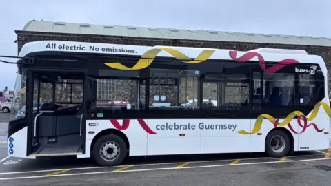 A white electric Stagecoach bus decorated with red and yellow ribbon graphics and the words "celebrate Guernsey". Text along the roofline reads "All electric. No emissions." The bus is parked outside a large stone building under a grey sky. 