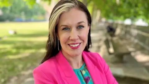 A woman in a pink blazer, brown and blonde hair, smiling at the camera in a park