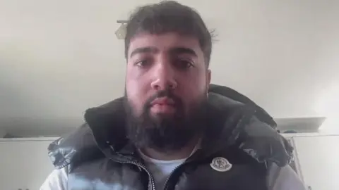 Zak Hanman has a dark beard and short dark hair. He is wearing a black coat.