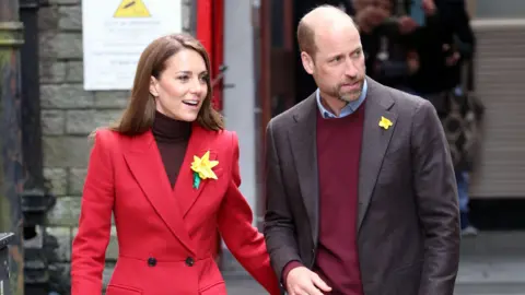 Getty Images Princess Kate and Prince William wearing formal attire with daffodils attached to their jackets. Kate is wearing a red coat with a turtleneck. He is wearing a suit jacket with a burgundy sweatshirt underneath.