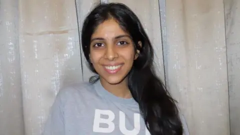 Aaliyah Patel Aaliyah Patel, with long black hair and a grey top, stands in front of a shimmering silver and cream curtain