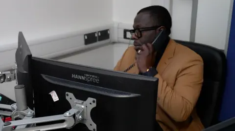 A man with cropped black hair sits at a desk in an office with white walls. He is speaking on the telephone. He is wearing a tan jacket and is positioned behind two computer monitors.