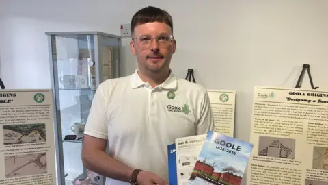 Stephen Walker stood in front of a display, smiling at the camera, holding leaflets about Goole. He has short brown hair and is wearing a white polo shirt.