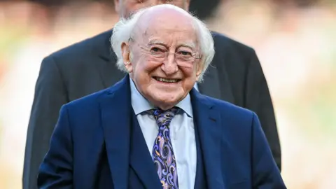 Getty Images Michael D Higgins, who has long, white hair smiles at the camera as he walks. He is wearing small, rimless glasses, a navy blue suit jacket and waistcoat, a lighter blue collared shirt and a patterned tie. There is a person standing behind him but he is blurred.