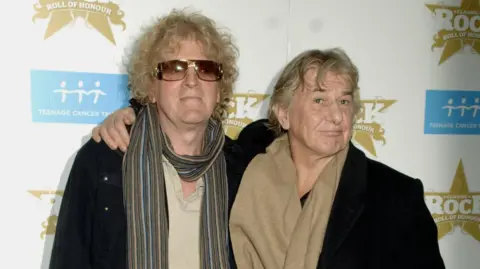 Ian Hunter (left) and Mick Ralphs (right) arrive for the Classic Rock Roll of Honour Awards in 2006. Hunter has curly blond hair and large sunglasses. He is wearing a striped scarf with a beige t-shirt and a black coat. Ralphs has long, wavy blond hair with a brown scarf and a black coat. 