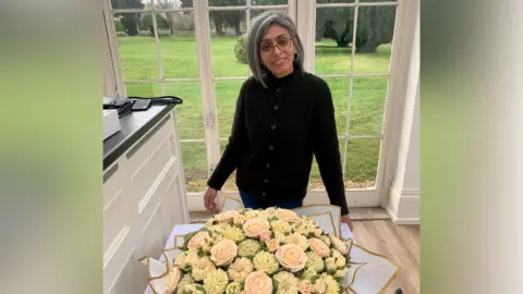 Ranj Patel A woman with shoulder-length silver hair and glasses smiles at the camera, in a kitchen in front of glass doors. On the table in front of her is an arrangement of floral cupcakes, designed to look like a bouquet.