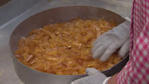 A copper pot on a silver slab full of orange Grays Herbal Tablets. You can see the hands of a worker. They are wearing grey gloves and a pink gingham apron. 