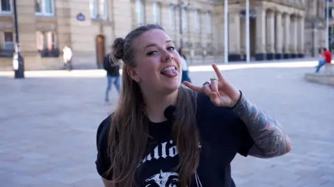 A woman with long hair, part of which is tied up in a bun, wears a black T-shirt with writing and a logo on. She is making a heavy metal sign with his index and little fingers. She has tongue and nose piercings and tattoos on her left arm