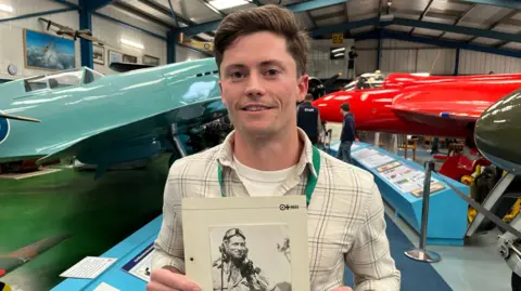 George Carden/BBC Michael McManus standing in one of the hangers holding a picture of his great uncle Ted Hall who is wearing a flying cap and goggles. Both are wearing shirts. Behind Michael is a sky blue Spitfire and red Hawker Hunter jet