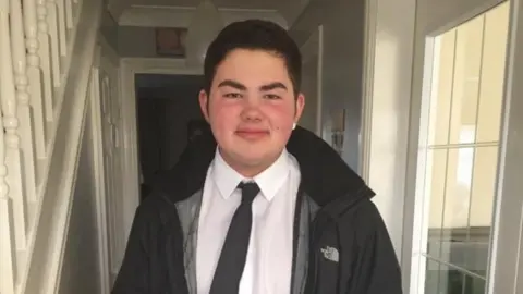 Family handout Andrew Rowlands. He is standing in the hallway of a house. He has black short hair and brown eyes. He is wearing a white shirt and black tie and a black coat. He is smiling at the camera.