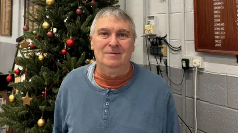 Gordon, a man with short hair, stands in front of Christmas tree wearing a blue button-up top