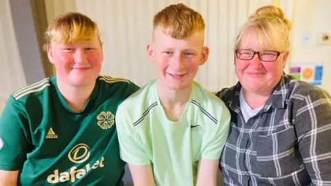 Adam Golebiewski in green tee shirt, in between sister Sophia in dark green football shirt, and mum Joanne in grey checked shirt, all smiling.