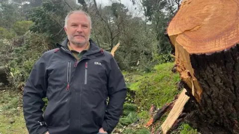 Darren Little is wearing a dark raincoat and is standing near a trunk of a tree. There is huge rip to the bark and a clean line where it has been cut through by a saw. this reveals a significant number of rings through the trunk, suggesting the tree was quite old. There is debris of other damaged trees behind him.