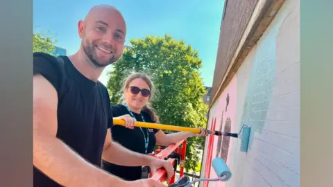 Luton Lifelines Harry and another woman stand on a platform holding paint rollers, which have blue paint on. They are close to the wall and are painting the mural, but look sideways at the camera and smile.
