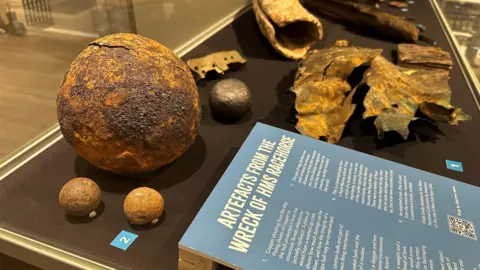 Items, including a large cannon ball, in a glass case along with a blue information board.