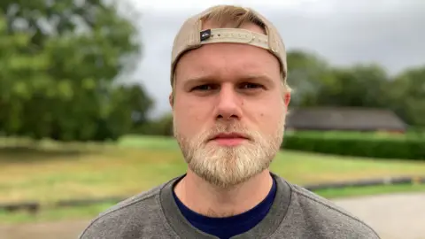 Harry Skinner looks directly at the camera. He has fair hair and a beard. He is wearing a back-to-front beige baseball cap and a grey sweatshirt over a navy blue T-shirt. He is outside and grass, trees and a building are visible behind him.