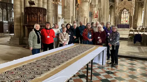 Tom Jackson/BBC A group of people standing under the cathedral's nave. The long piece of embroidery is placed in front of them.