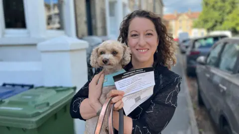 Donna Ferguson holding her small dog in her arms and a flier saying "Cambridge needs fairer HMO controls". Donna has curly brunette hair and is wearing a black top. She is stood on a street pavement with houses on one side and cars to her right.
