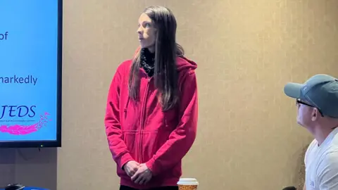 Amy is in a red hoodie stands near a large presentation screen displaying text and the JEDS logo. Another person seated nearby in a white T‑shirt and cap looks toward the presenter. Papers, drinks, and a laptop are on the tables in front of them.