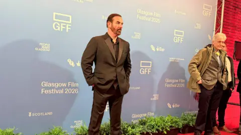 A man in a black suit stands in front of a blue background