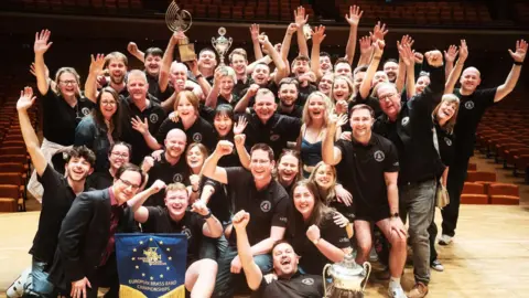 Approximately fifty people are gathered as a group on a stage in the foreground with theatre seating in the background. The members of Flowers Band are of various ages and are all looking at the camera. Some people have their hands open in the air, some are fist pumping the air, others hold aloft trophies. They are all wearing black polo shirts with the band's logo on except composer Oliver Waespi who is wearing a burgundy button-up shirt with a black blazer, dark short hair and dark rimmed glasses.