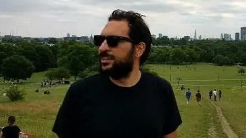 University of St Andrews Dr Antonis Vradis is standing outdoors and there is a large green open space behind him. He has dark hair, is wearing sunglasses and a black T-shirt. 