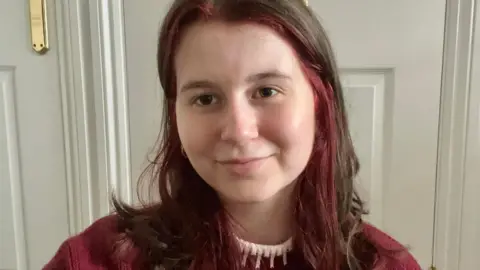Abi Wilson Selfie photograph of Abi Wilson, 20, who has brown and red hair. She stands in front of a white door frame and smiles at the camera. She has brown eyes and wears a red jumper with a white trim collar. 