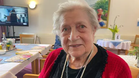 Current Ilford Park resident Alicia Rosanna. She has short grey hair and is wearing a red cardigan and black shirt. She is wearing pearls. 