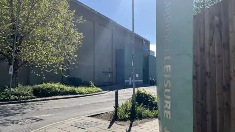 Phil Shepka/BBC A side entrance to Cambridge Leisure Park, showing the side of the big brick building housing the cinema. There is greenery and a road. It is a sunny evening and there is some lens flare on the image.