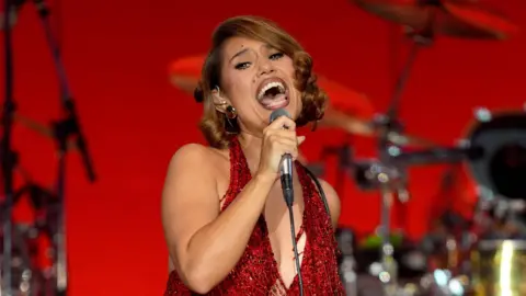 PA Media Singer Raye is pictured singing at a gig. She is wearing a red dress. She has her light brown hair worn in a bob.