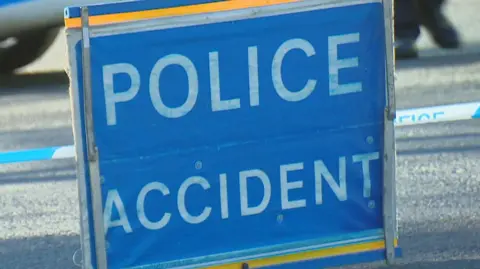 BBC Police accident sign, white block capitals on blue sign, with blue and white police tape.