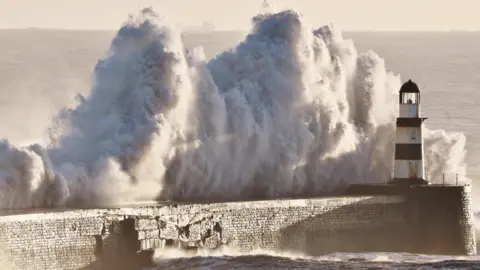 BBC Weather Watchers/Dan Seaham Harbour Lad Tall white, foamy ocean waves are about to crash over a pier with a black and white lighthouse at the end of it. The waves are almost twice as high as the lighthouse.