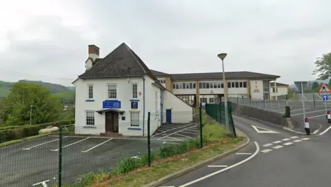 Google Wide shot of Ysgol Bro Caereinion. A white school house can be seen on the left hand side, with a much bigger, modern school building behind it. A road runs underneath a second floor bridge and a school yard can be seen on the right hand side. 