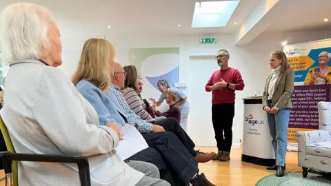 A group of older people sitting in a row on chairs, listening to Simon Allen who is standing up addressing them in a red jumper with Wera Hobhouse standing to his side in a smart jacket and jeans