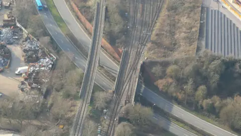 Network Rail An aerial view of a railway bridge over a dual carriageway road. There is a waste yard on the bottom left of the picture.