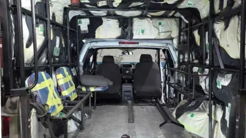 A vehicle, with folded-up seats in it, with items stuffed into the walls. You can see two seats at the front and the vehicle is mostly empty. 