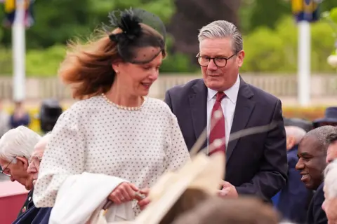 PA Media Prime Minister Sir Keir Starmer and his wife, Lady Victoria, seek their seats. She is wearing a white dress and he wears a dark suit and red tie.