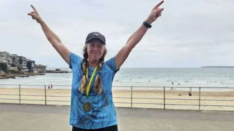 PA Media Nikki Love's arms are raised in a victory pose at Bondi Beach. She has long, braided, fair hair and wears a baseball cap, a blue sports top and a medal around her neck.