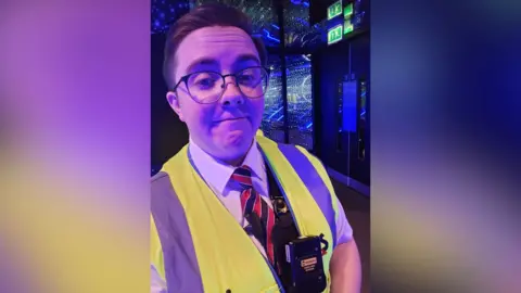 Lyndsey Higgins Lyndsey Higgins wears a white shirt with a black-and-red striped tie underneath a yellow high-viz vest. There is a body-worn camera attached to the vest. 