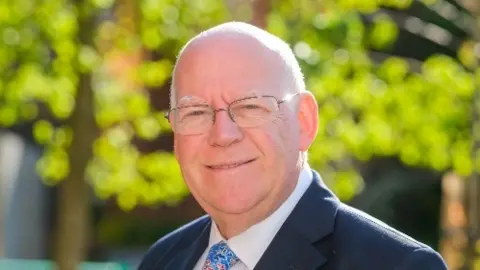 A picture of Jeff Brooks, who is wearing a dark suit and is standing in front of a tree, which is blurred in the background. He is wearing glasses and is bald or has very short white hair.
