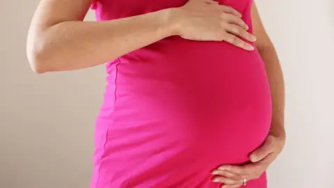 The torso of a woman wearing a bright pink dress who is eight months pregnant. She has her hands on the top and bottom of her baby bump.