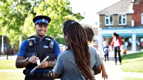 Reading Borough Council A police officer smiling while talking to a man who appears to be holding a child. A woman is seen reaching out for the hand of her child in the background. It is a sunny day.