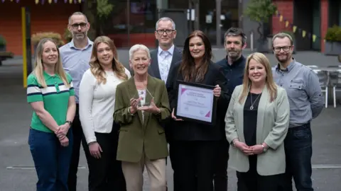 Anglia Ruskin University Nine people are standing in a group facing forwards. Two women, who are in the middle, are holding awards. They are standing in a courtyard which has seating.