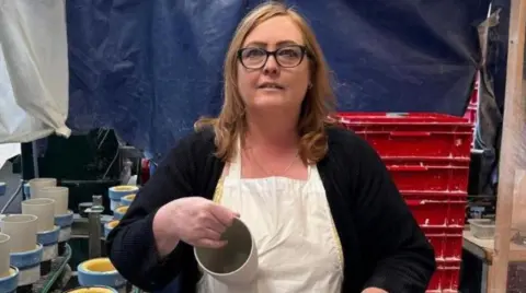 Sharon Yates is looking at the camera, and has shoulder-length brown hair and has black-framed glasses on. She is sat down and has a white apron on over a yellow top and a dark cardigan. In her hand is a small pot, and behind her is a pottery production area.