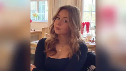 An image of Jessica Glasser sitting at a table. She is wearing a long sleeve black top and gold necklaces. She has long, waved blonde hair and is smiling at the camera.