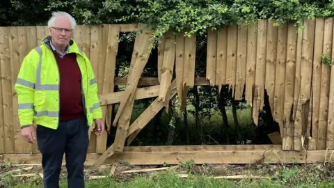 Amy Holmes/BBC A man in a high-visibility jacket stands beside a wooden fence with a large section smashed through, showing extensive impact damage and broken panels.