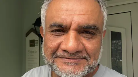 Gulam Patel has short grey hair and a short grey beard, he wears a grey t-shirt